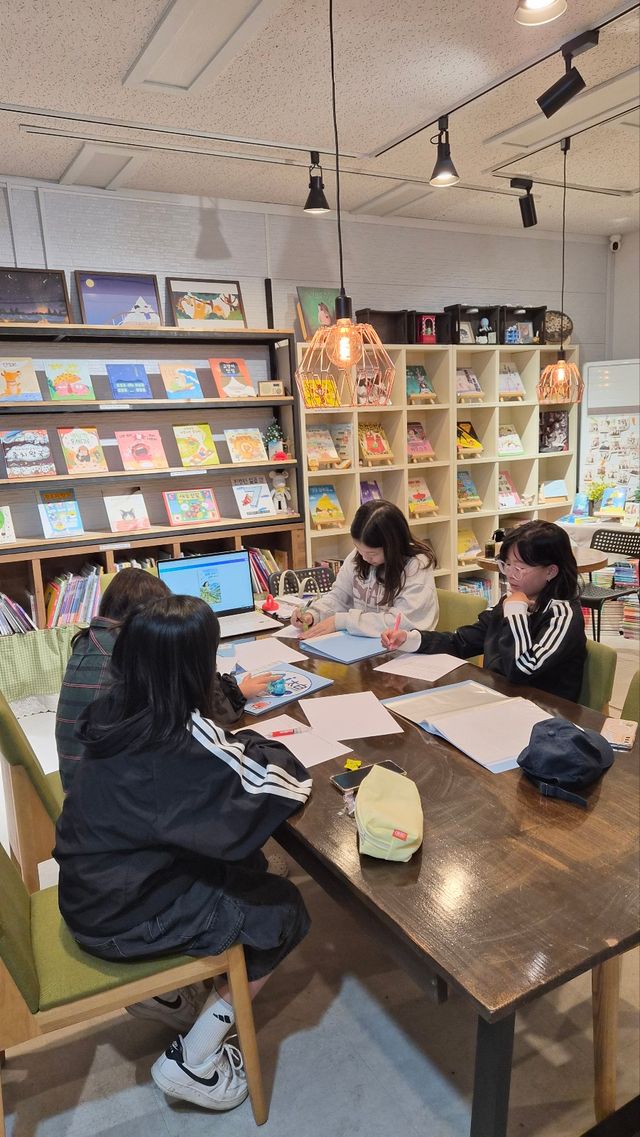 그림책이 진열된 실내 공간에서 아이 네 명이 테이블에 앉아 활동지를 쓰고 있다. 노트북 화면이 보인다.