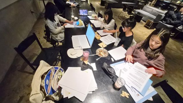 어두운 카페의 긴 테이블에서 아이들이 노트북과 종이 자료로 활동하는 모습.